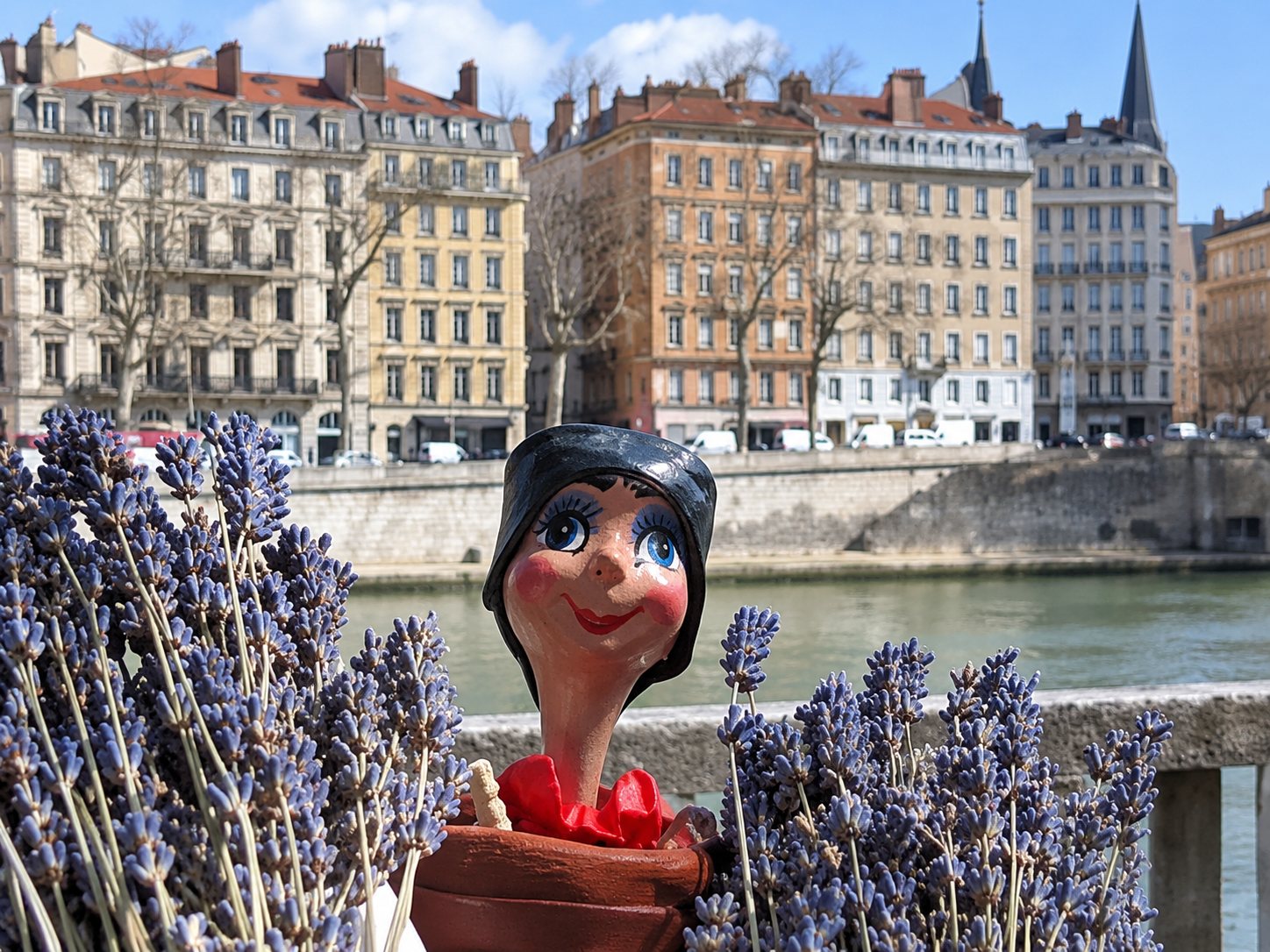 Une marionnette à écharpe rouge se tient entre deux bouquets de lavande, avec en arrière-plan un fleuve et des immeubles anciens sous un ciel bleu.
